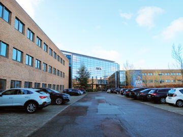 Facade af en moderne kontorbygning med store glaspartier og parkerede biler foran.