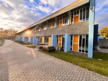 Udendørsbillede af Skaarup Skole, en to-etagers bygning med blå og orange detaljer, sollys og flisebelagt sti.